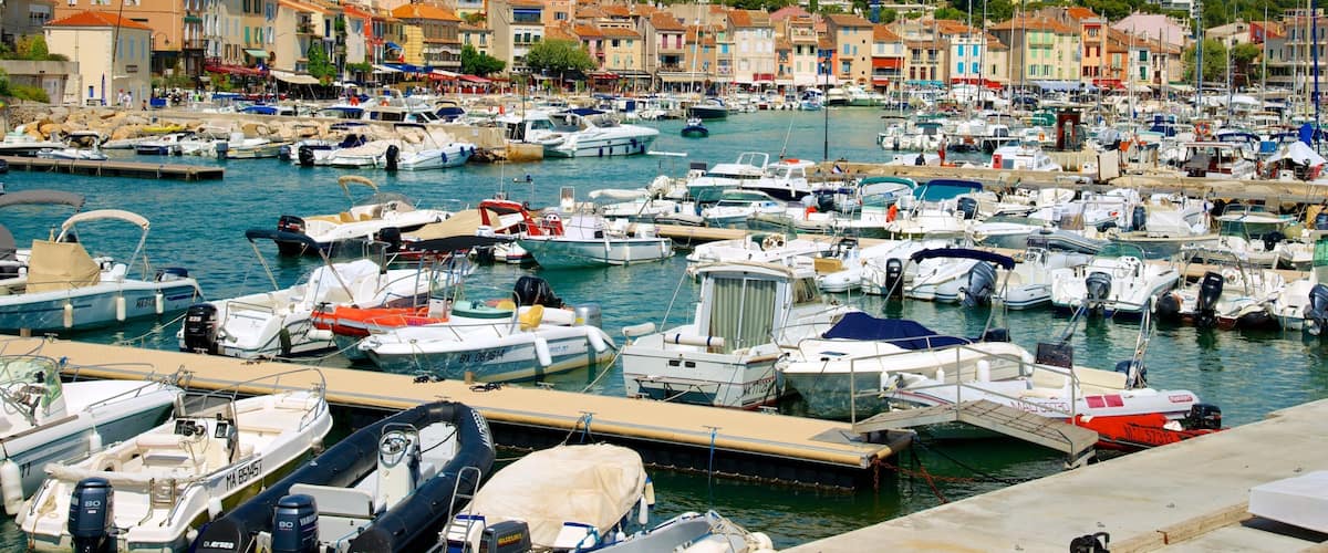 A marina filled with various boats docked in calm waters, surrounded by colorful buildings and a hillside in the background.