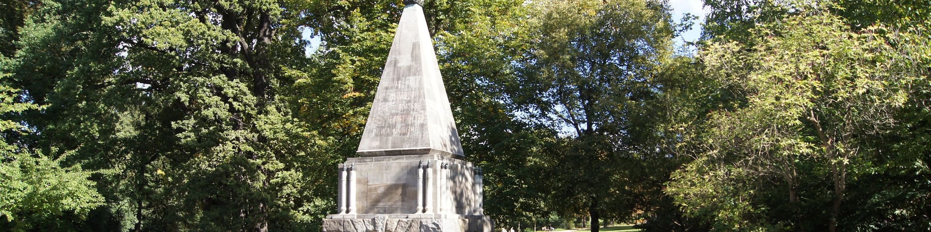 A stone obelisk monument surrounded by trees and a paved area.