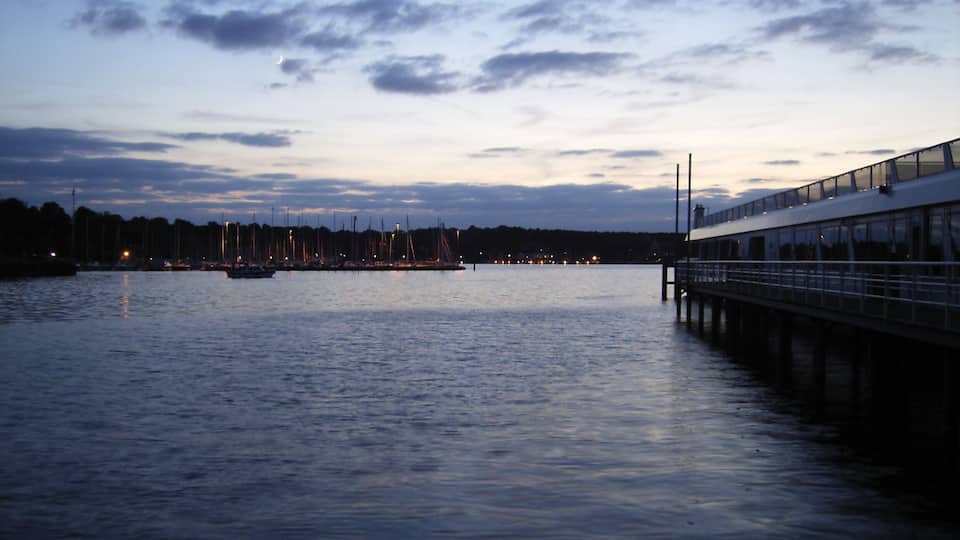 Um areal calmo ao entardecer com um cais que se estende na água, barcos atracados e a silhueta de uma cidade ao fundo.