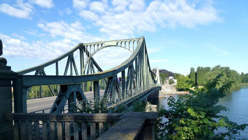 Uma ponte de aço com design curvo, um rio abaixo e vegetação nas margens.