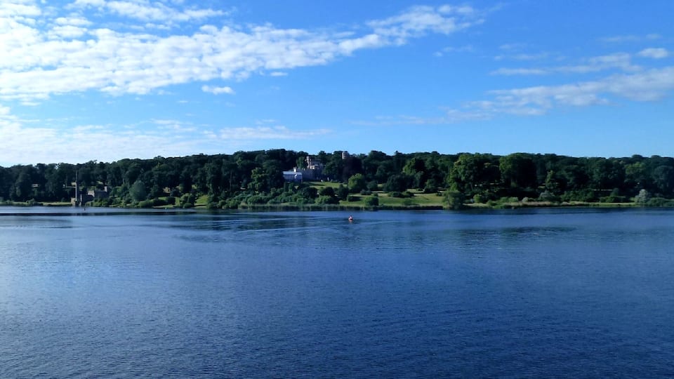 Um lago calmo com uma pequena ilha ao centro, cercado por vegetação exuberante e um céu azul límpido.
