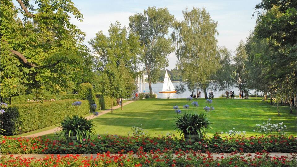 Um parque com gramado, canteiros de flores e um veleiro na água.