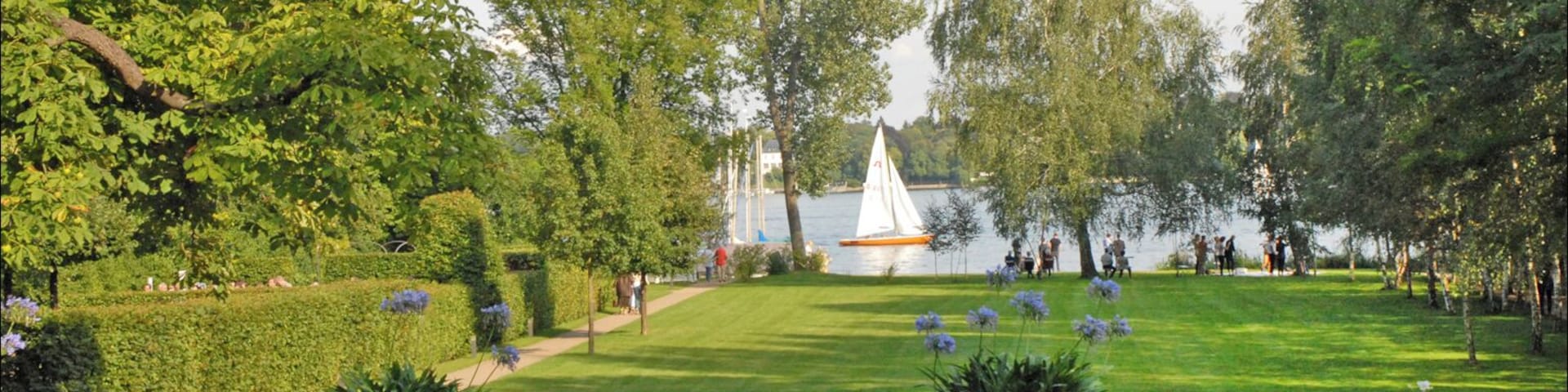 Un parque con césped, macizos de flores y un velero en el agua.
