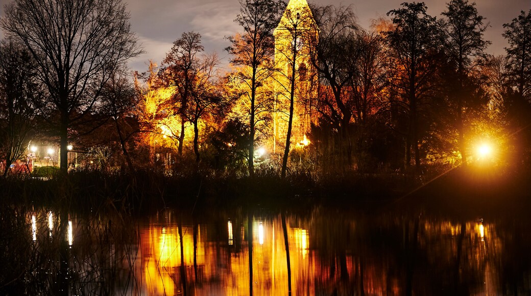 Une église illuminée, surmontée d’une croix, entourée d’arbres et dont le reflet se mire dans une étendue d’eau.