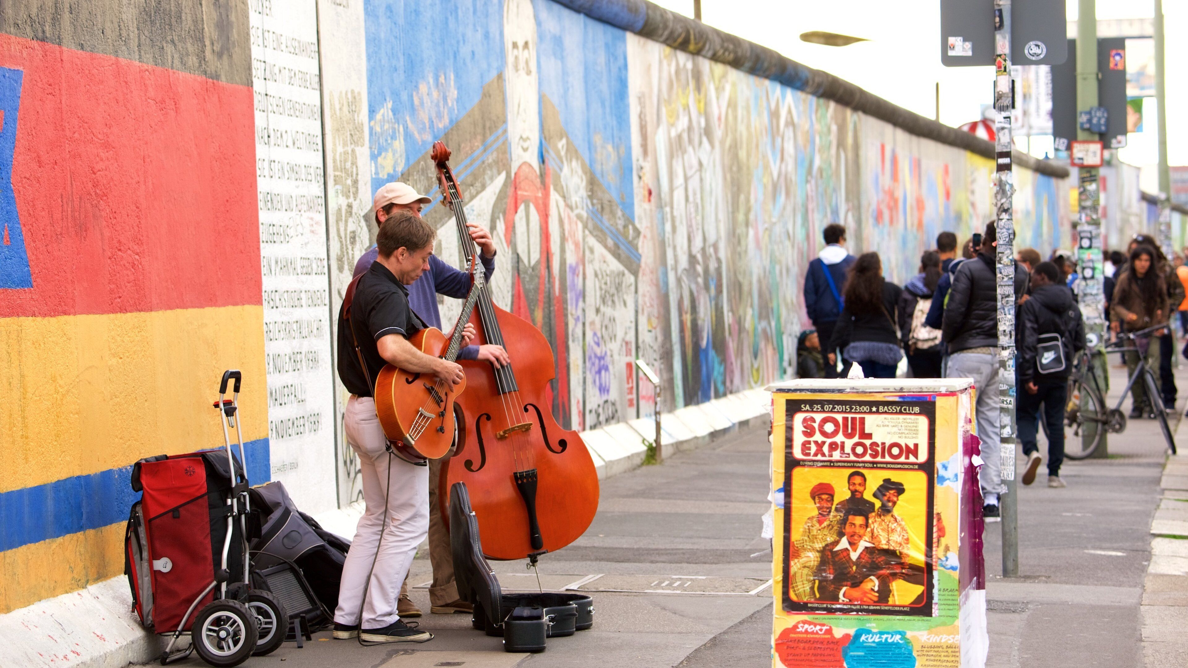 A street musician playing a double bass and a guitar, with a poster for an event in the foreground.