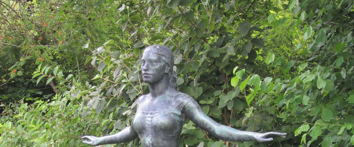 A bronze statue of a woman in a skirt and dress, standing on a pedestal in a garden with lush green foliage in the background.