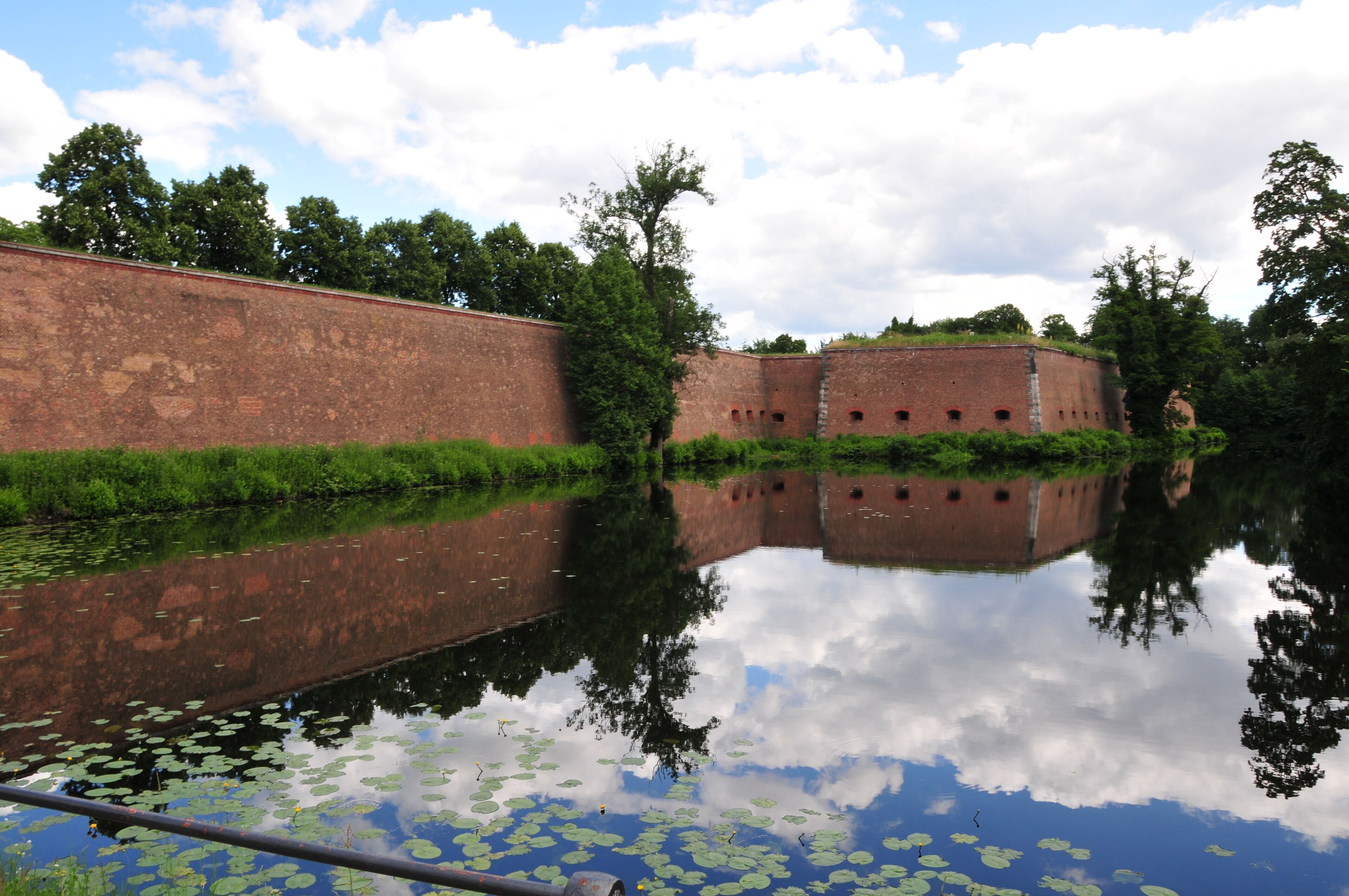 Das Bild zeigt die rechte Seite vom Haupttor der Spandauer Zitadelle in Berlin Spandau, Deutschland. Zu sehen ist auch etwas vom Zitadellengraben bei Windstille an einem sonnigen Tag.