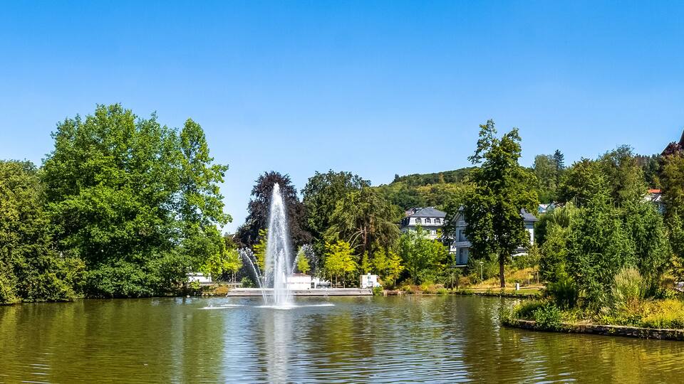 A serene lake with a central fountain, surrounded by lush greenery and residential buildings.