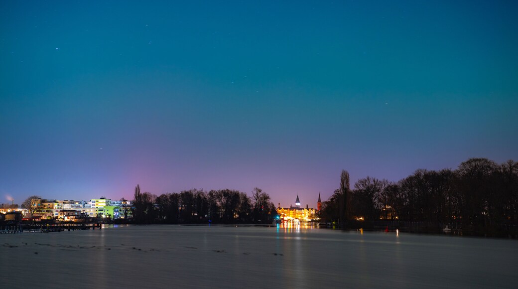 Nachtszene mit ruhigem Gewässer, das Stadtlichter reflektiert, einer Skyline mit beleuchteten Gebäuden und klarem Himmel.