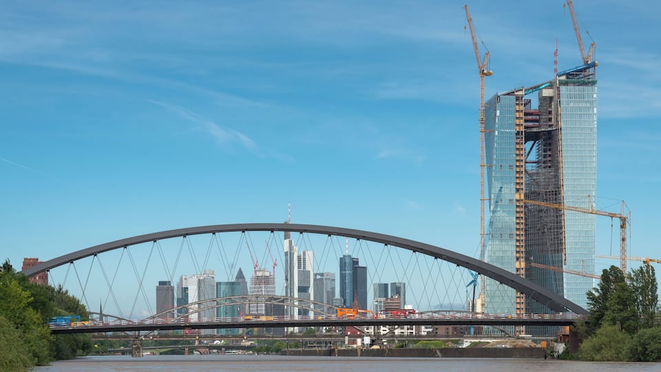 Un pont enjambant une rivière, avec la silhouette d’une ville en arrière-plan et un grand immeuble en construction.