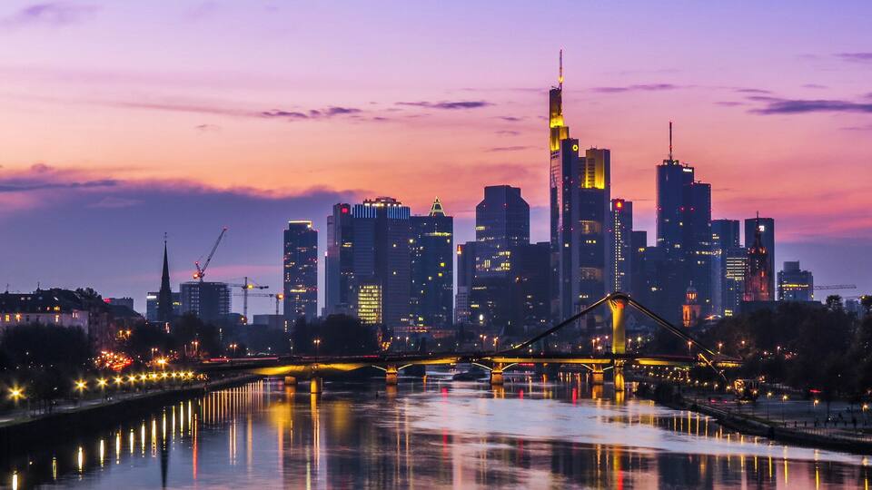 Een stadszicht in de schemering met een rivier die lichtjes weerkaatst, een opvallende brug en meerdere wolkenkrabbers.