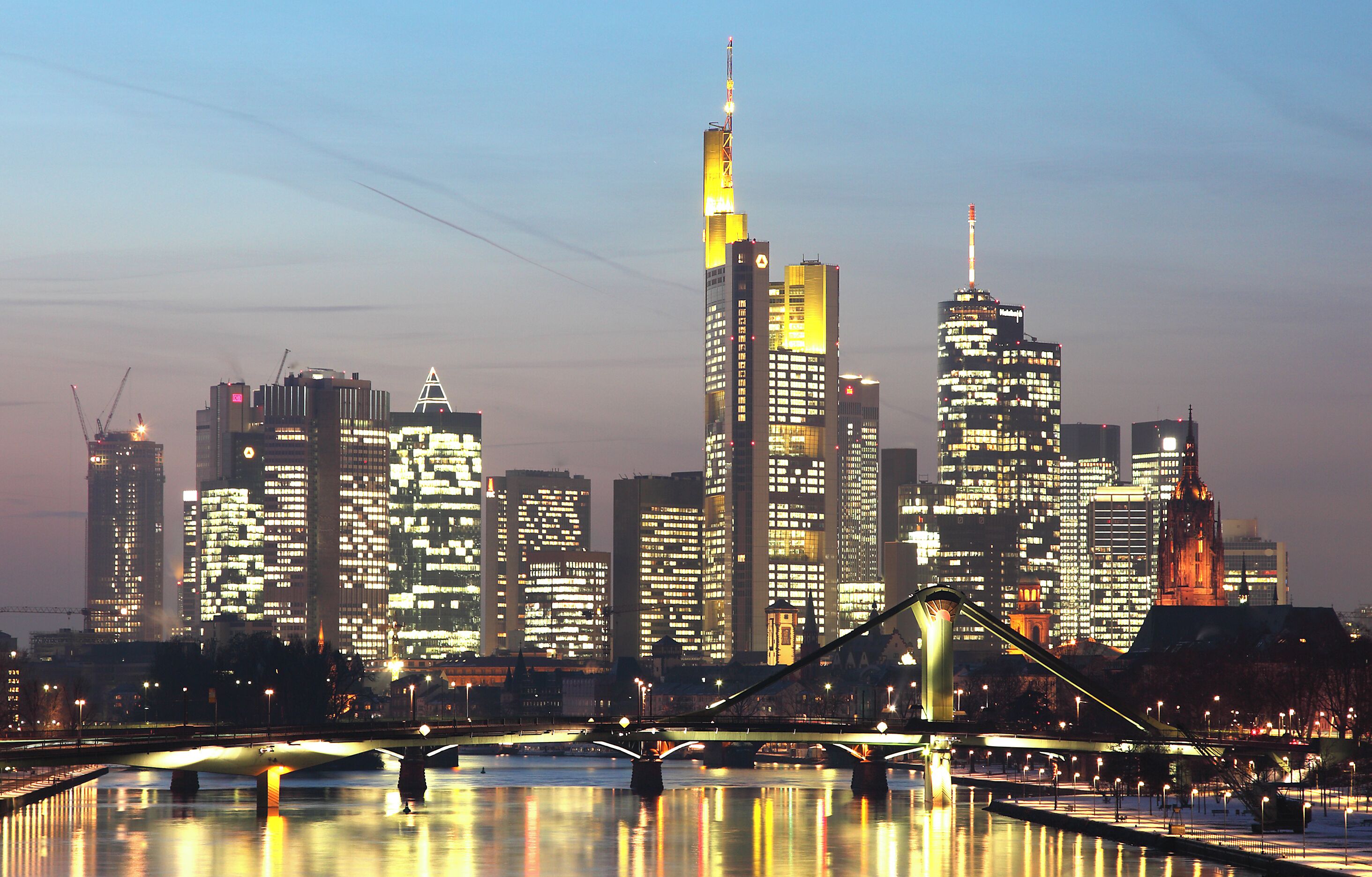 Une vue sur les gratte-ciel d’une ville au crépuscule, avec des bâtiments illuminés et un pont enjambant une étendue d’eau.