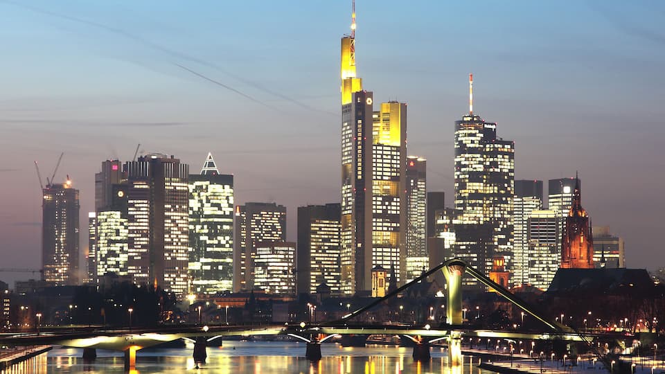 Une vue sur les gratte-ciel d’une ville au crépuscule, avec des bâtiments illuminés et un pont enjambant une étendue d’eau.