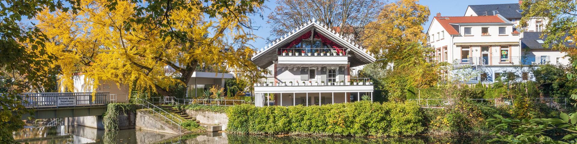 A building with a white facade and red roof, surrounded by autumn-colored trees and a calm body of water reflecting the structure.
