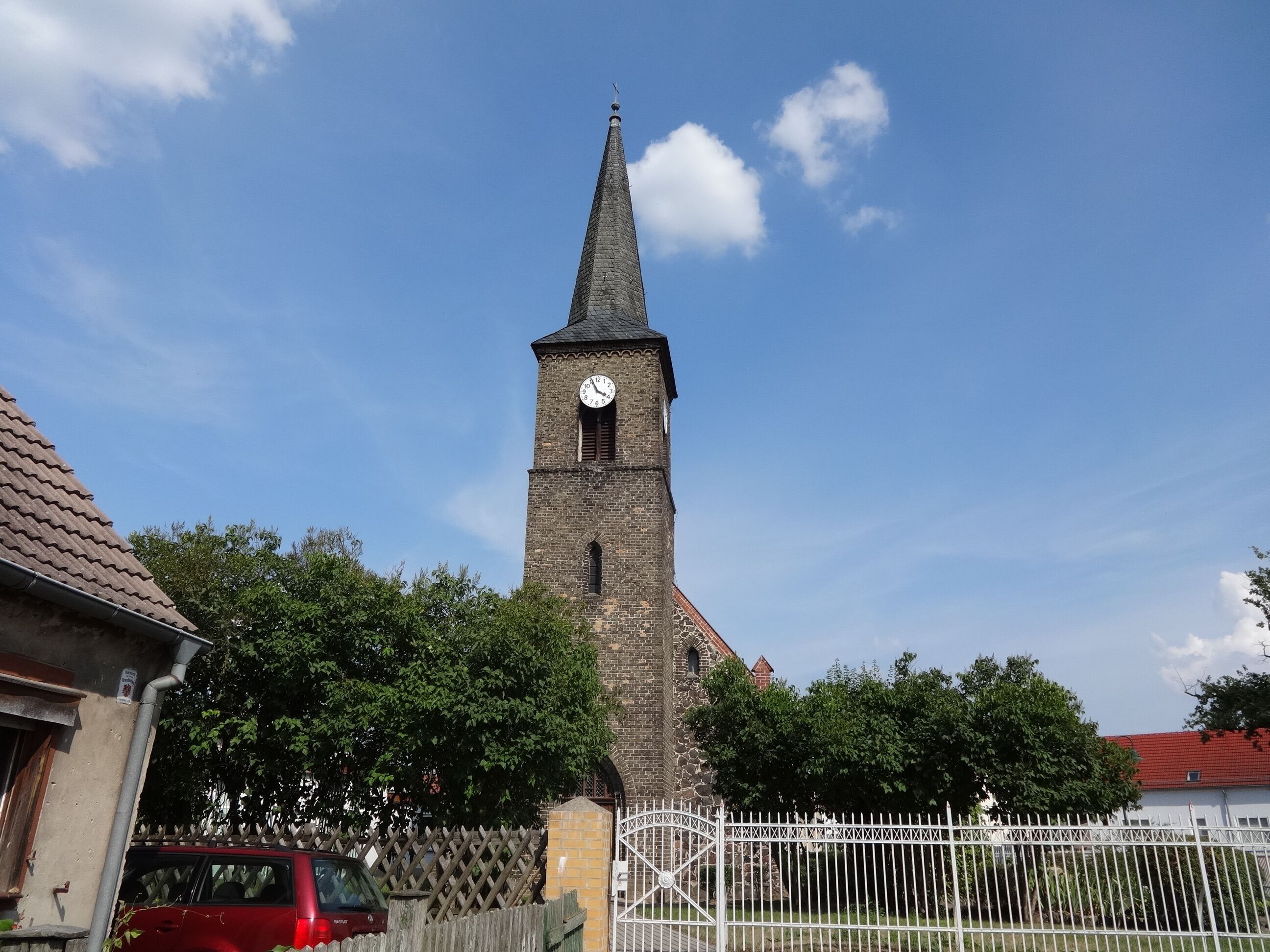 A tall church tower with a clock, surrounded by trees and a fence.