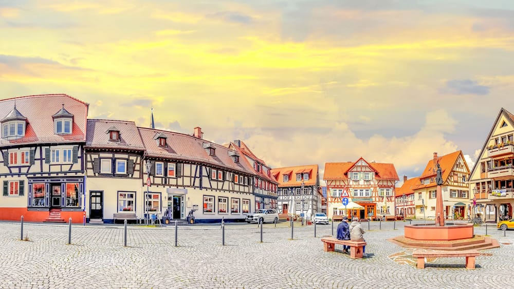 A cobblestone square with traditional half-timbered buildings, a central fountain, and a clear sky.