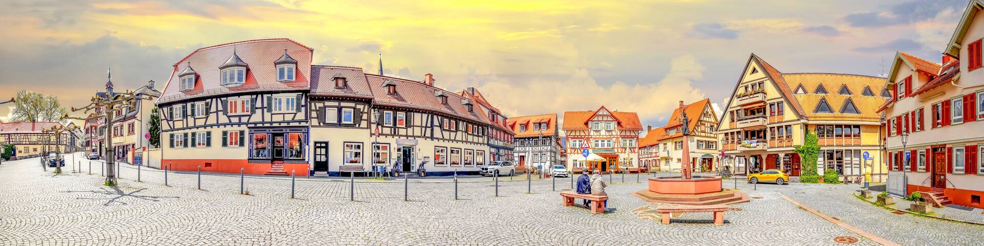 A cobblestone square with traditional half-timbered buildings, a central fountain, and a clear sky.