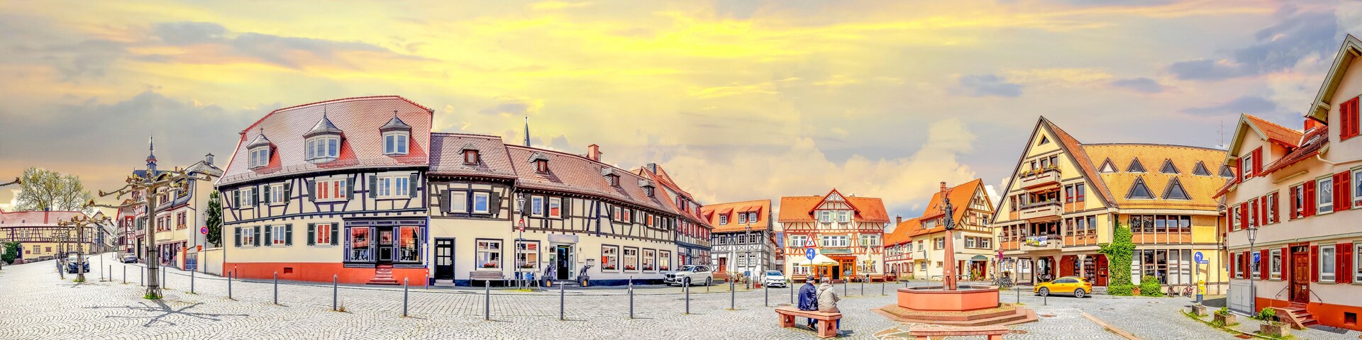 A cobblestone square with traditional half-timbered buildings, a central fountain, and a clear sky.