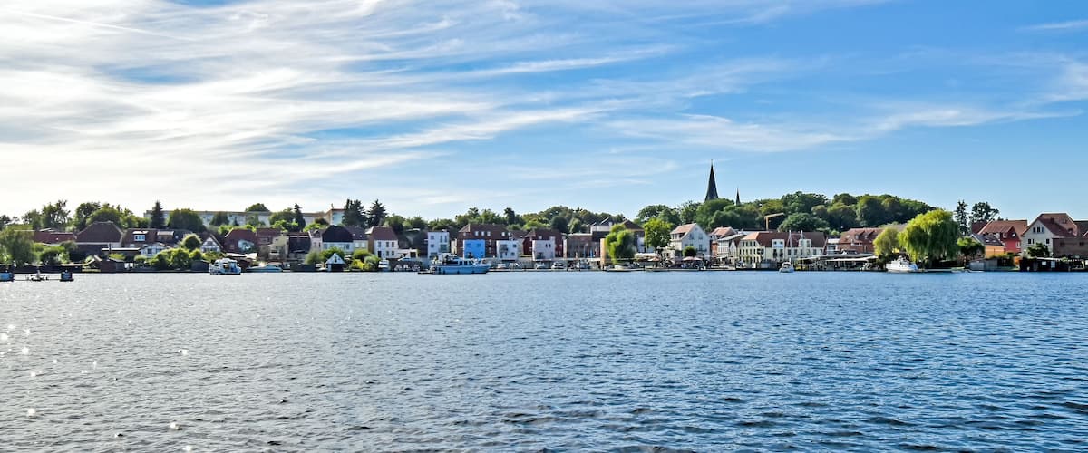 A riverside town with historic buildings, a church spire, and boats on the water.
