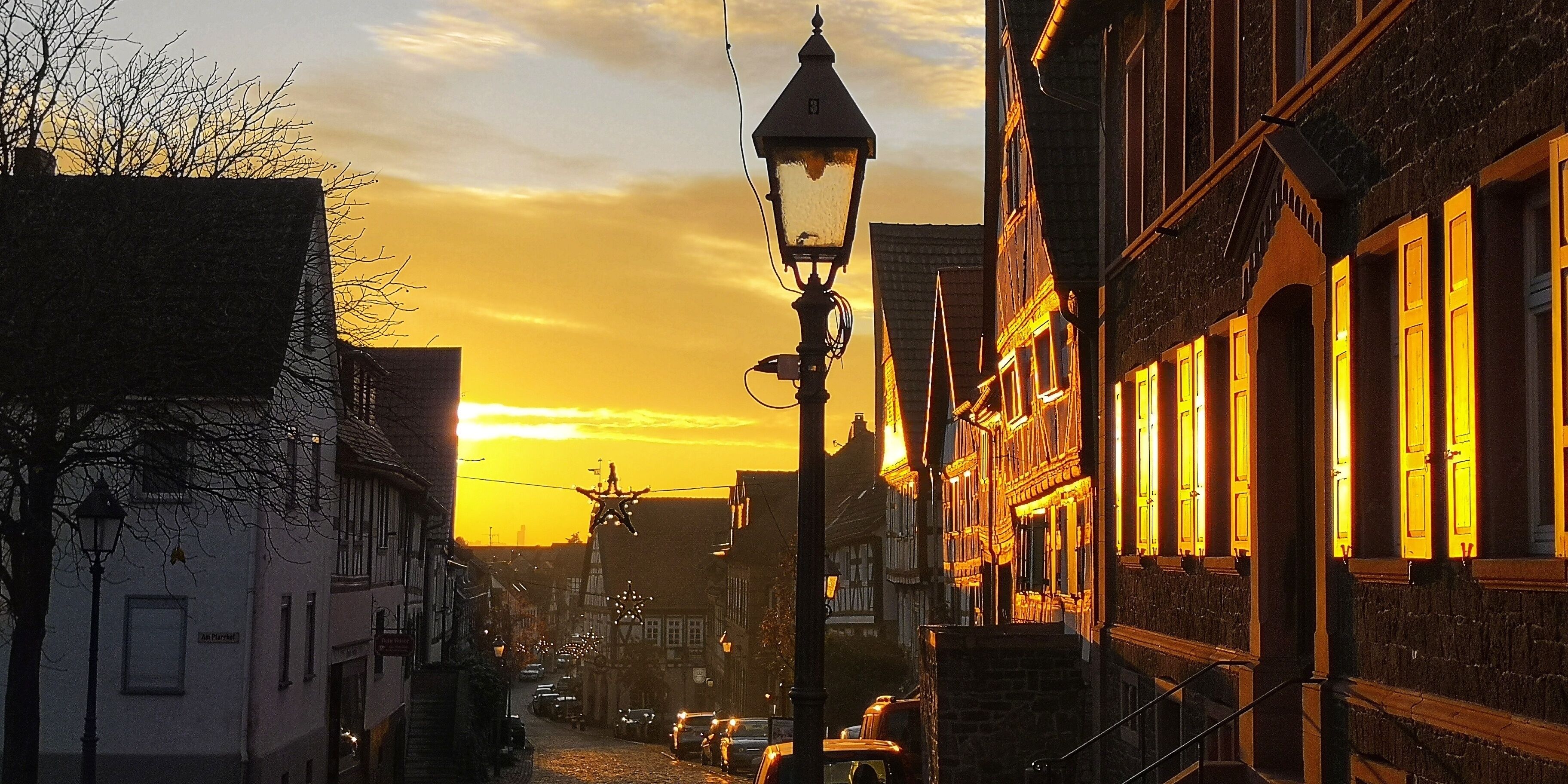 A cobblestone street lined with traditional half-timbered houses, a street lamp, and a star-shaped decoration against a sunset sky.