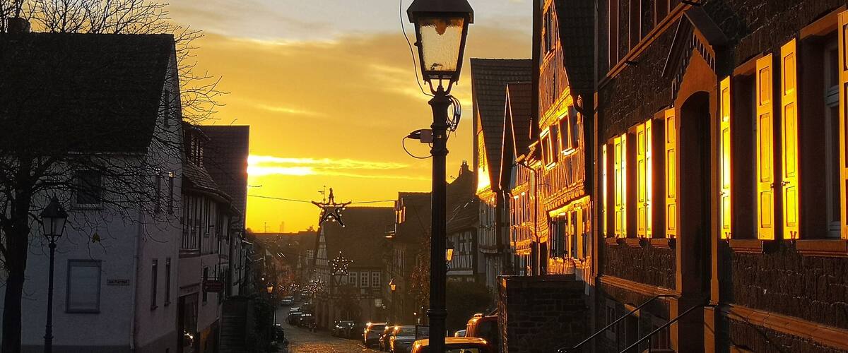 A cobblestone street lined with traditional half-timbered houses, a street lamp, and a star-shaped decoration against a sunset sky.
