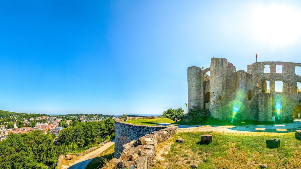 Eine historische Burgruine aus Stein mit einem Burggraben, umgeben von üppiger Vegetation und einer Stadt im Hintergrund.