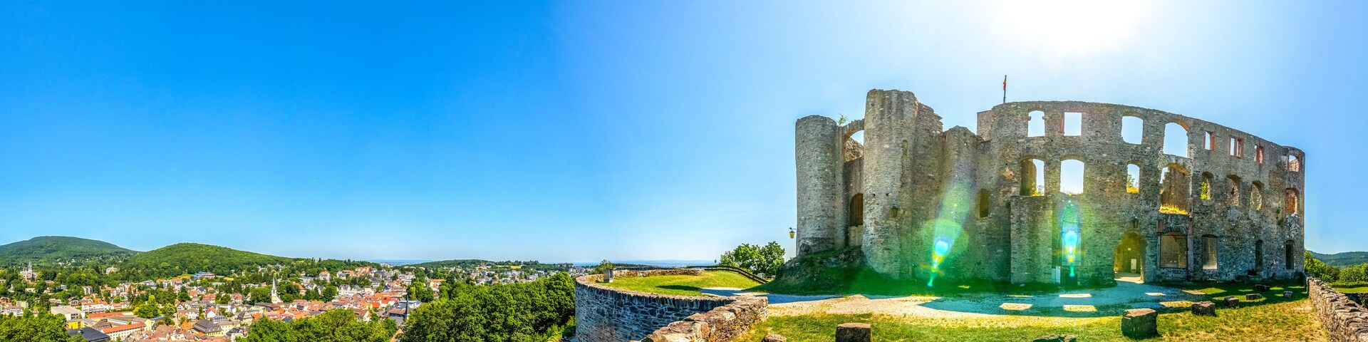 Ruinas de un castillo de piedra histórico con un foso, rodeado de abundante vegetación y un pueblo al fondo.