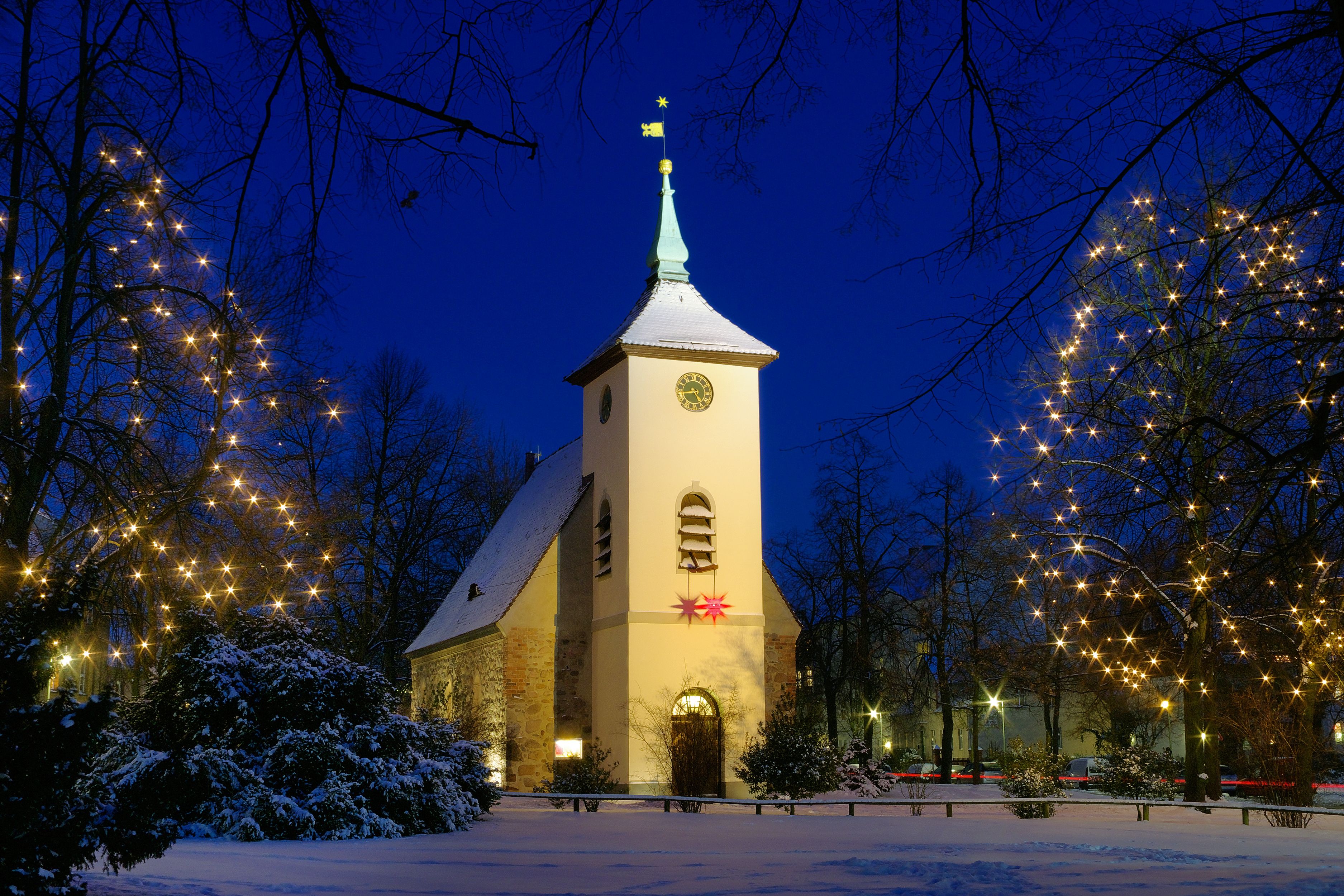 Una iglesia nevada con una torre de reloj, rodeada de árboles y decorada con luces en forma de estrella.
