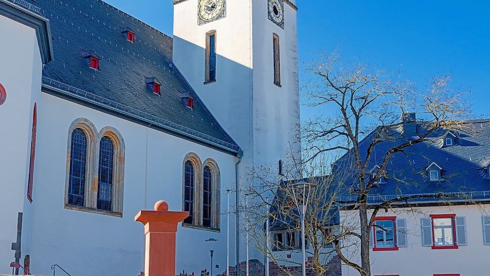 A church with a tall tower and a clock, a stone well, and a building with red window frames.