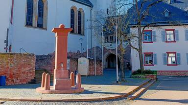 Une église dotée d’un haut clocher et d’une horloge, un puits en pierre et un bâtiment aux encadrements de fenêtres rouges.