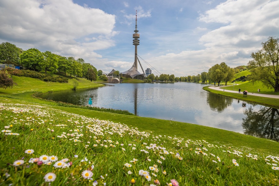 Ein See mit einem Turm im Hintergrund, umgeben von grünem Gras und Blumen.