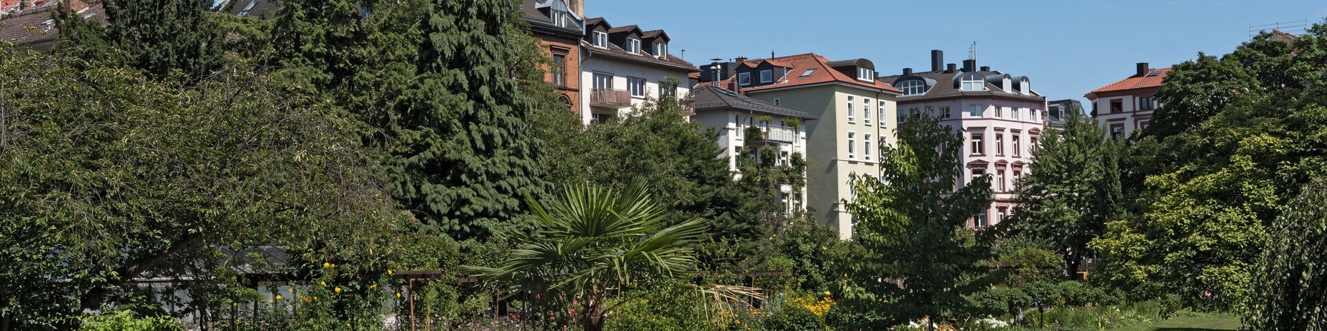 Un parque con un gran macetero de piedra, abundante vegetación y una hilera de edificios residenciales al fondo.