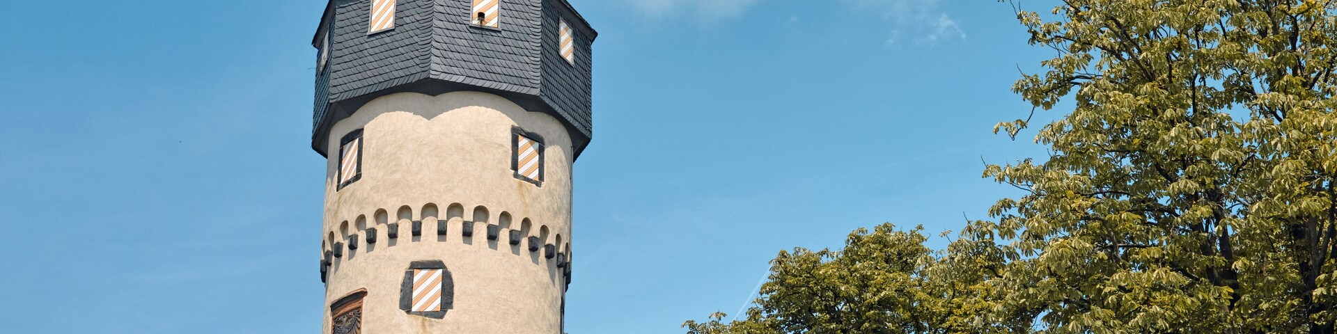 Una torre histórica con techo puntiagudo y veleta, rodeada de árboles y un cielo despejado.