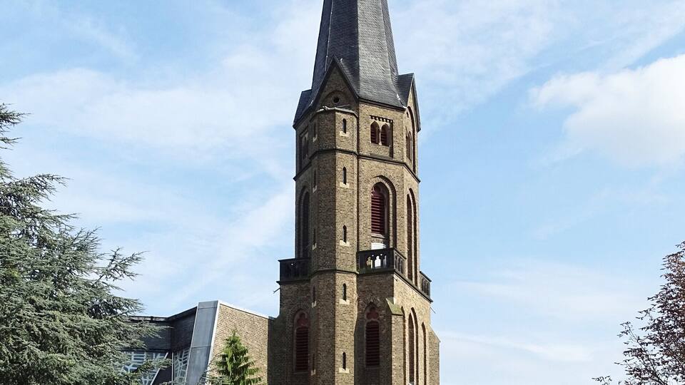 A tall church tower with a pointed spire, surrounded by trees and modern buildings.