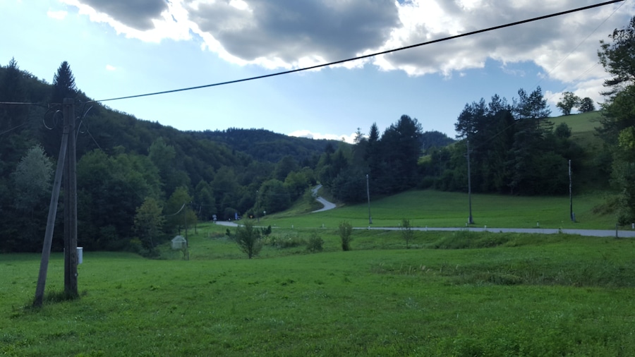 Una valle verde con una strada tortuosa, linee elettriche e una collina boscosa sullo sfondo.