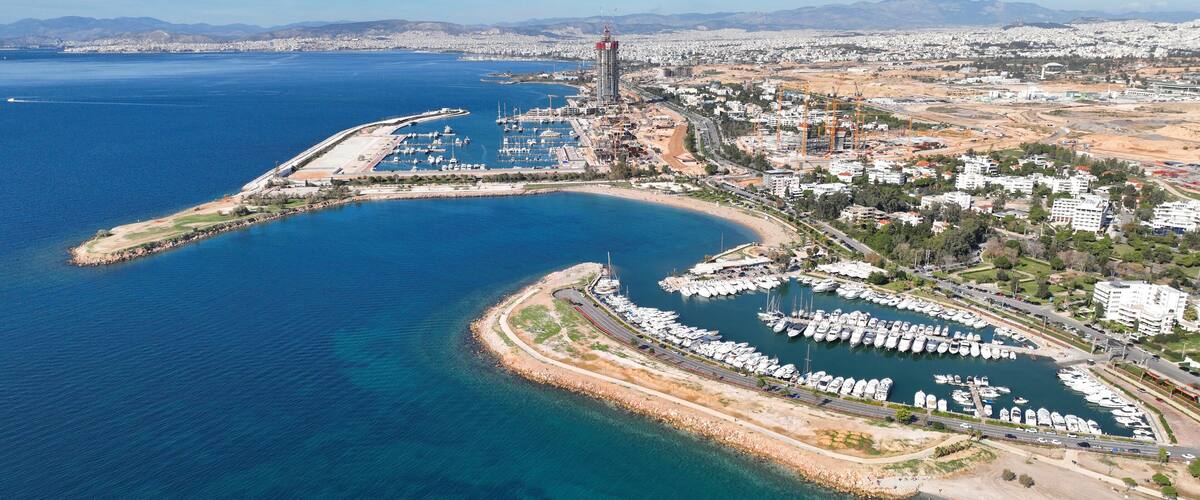 An aerial view of a coastal marina with boats, a large building under construction, and a cityscape in the background.