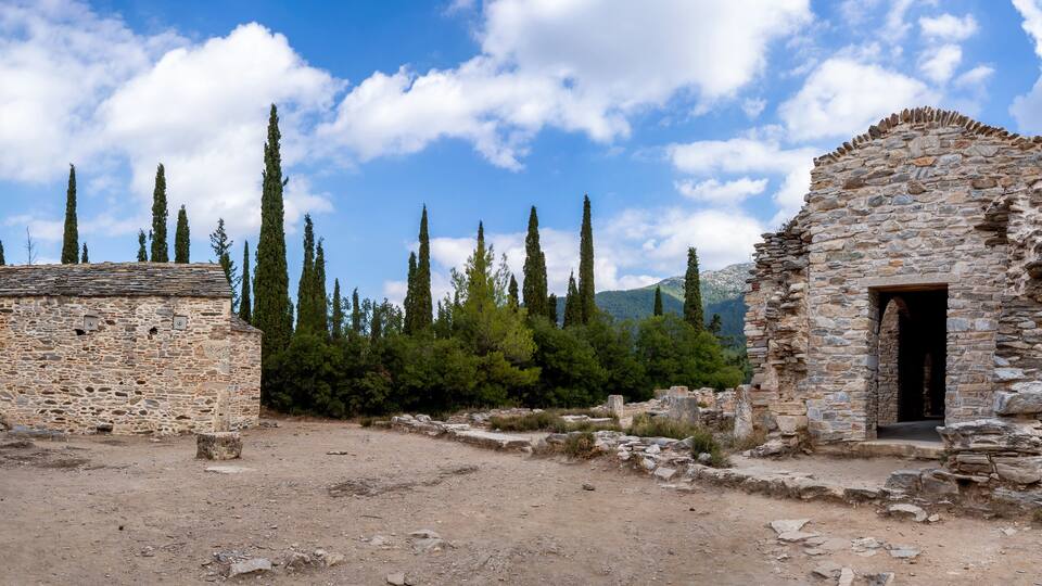 An ancient stone structure with a stone door and a small building with a tiled roof, surrounded by trees and mountains.