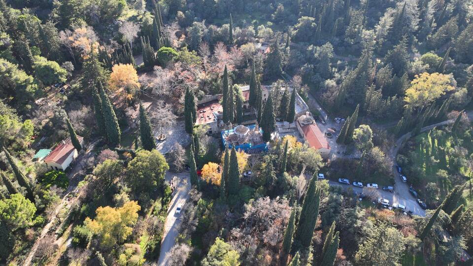 An aerial view of a forested area with a traditional building with a tiled roof, surrounded by tall trees and a winding road.