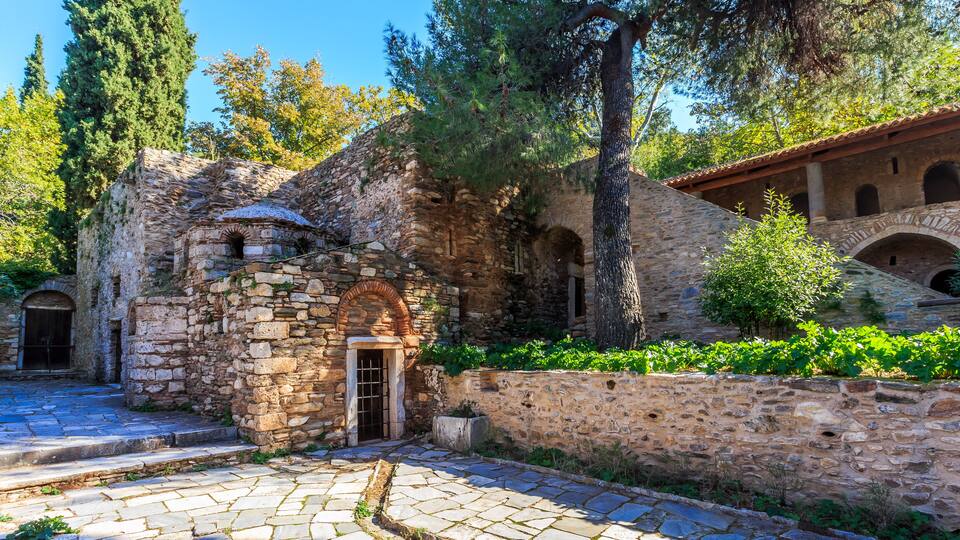 An ancient stone building with a wooden door, surrounded by trees and a stone wall.