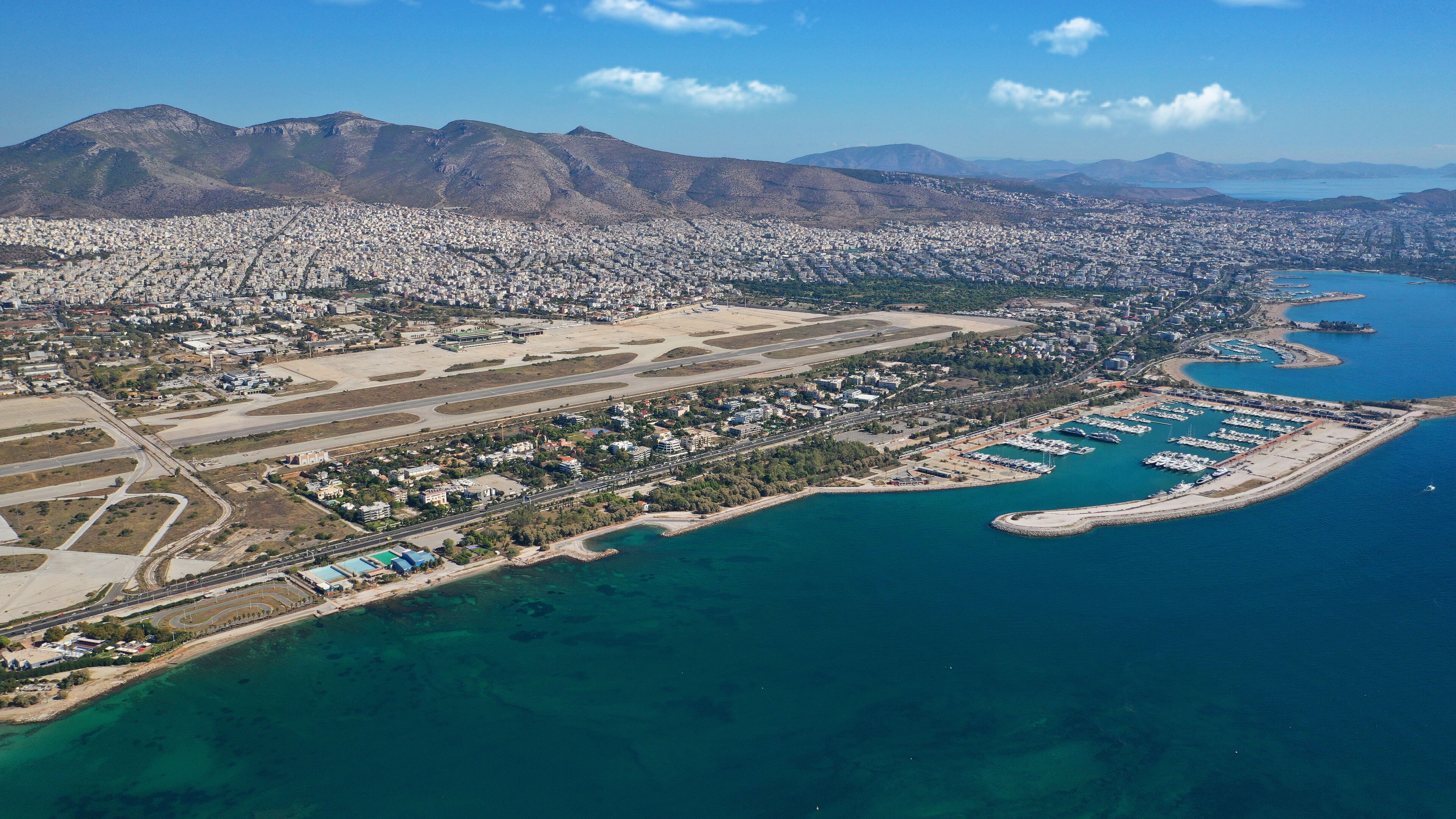 An aerial view of a coastal city with a marina, mountains in the background, and a large airport.