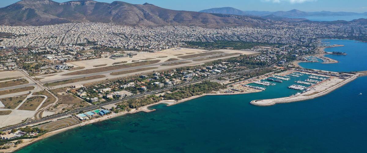 An aerial view of a coastal city with a marina, mountains in the background, and a large airport.