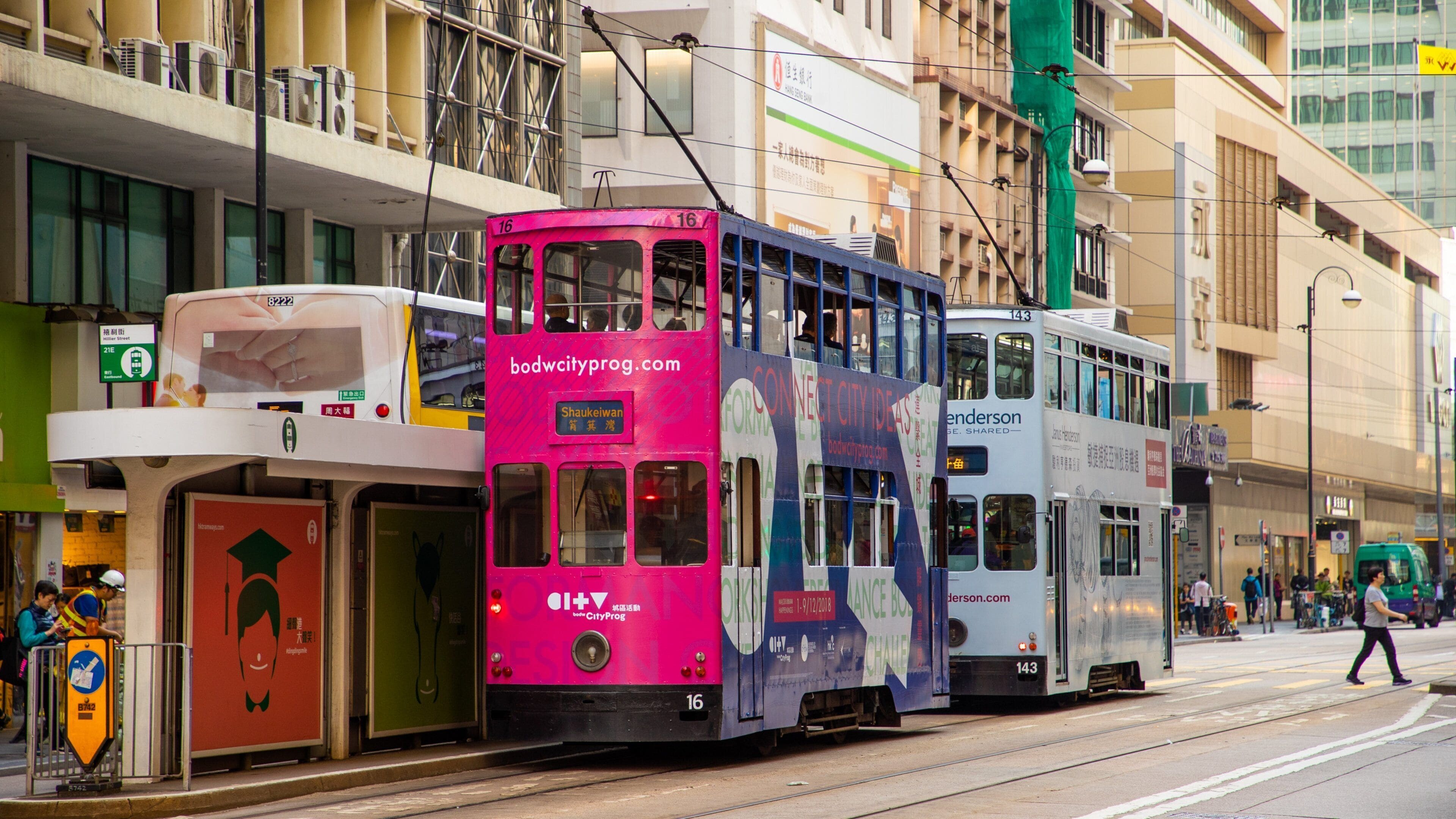 A pink and blue double-decker bus on a city street with buildings and pedestrians.