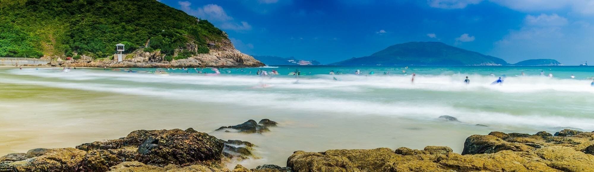 Una playa con aguas turquesas cristalinas, primer plano rocoso y montañas al fondo.