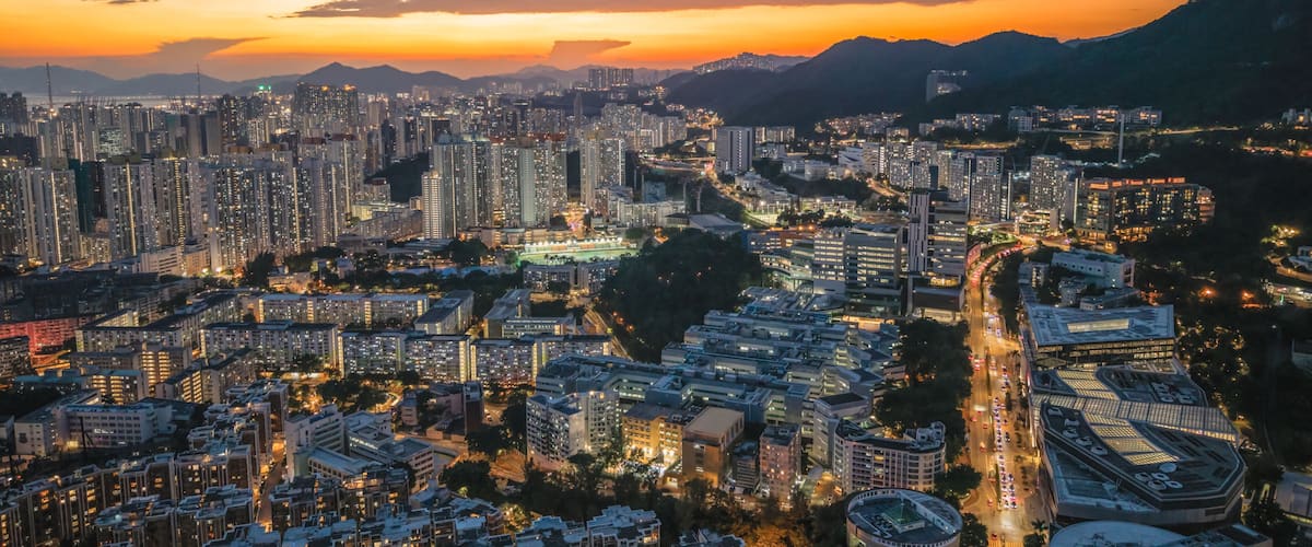 A cityscape at dusk with high-rise buildings, winding roads, and a mountainous backdrop.