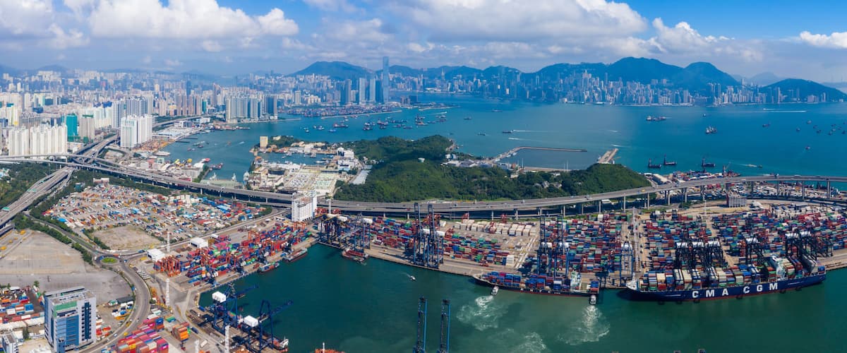 A bustling port with numerous cargo containers, a bridge, and a city skyline in the background.