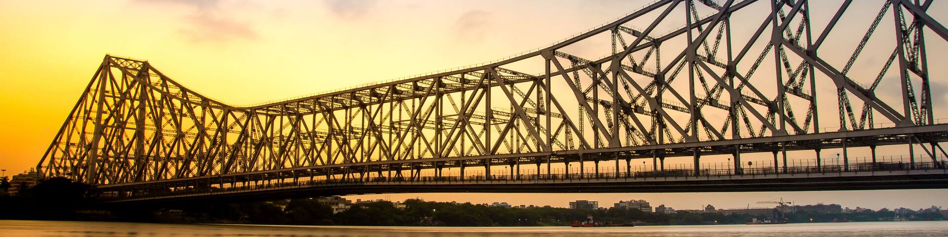 A large steel bridge spanning a river with a sunset in the background.