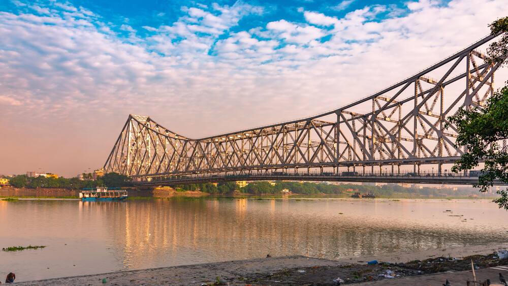 A large steel bridge spanning a river with a boat in the foreground and a cityscape in the background.