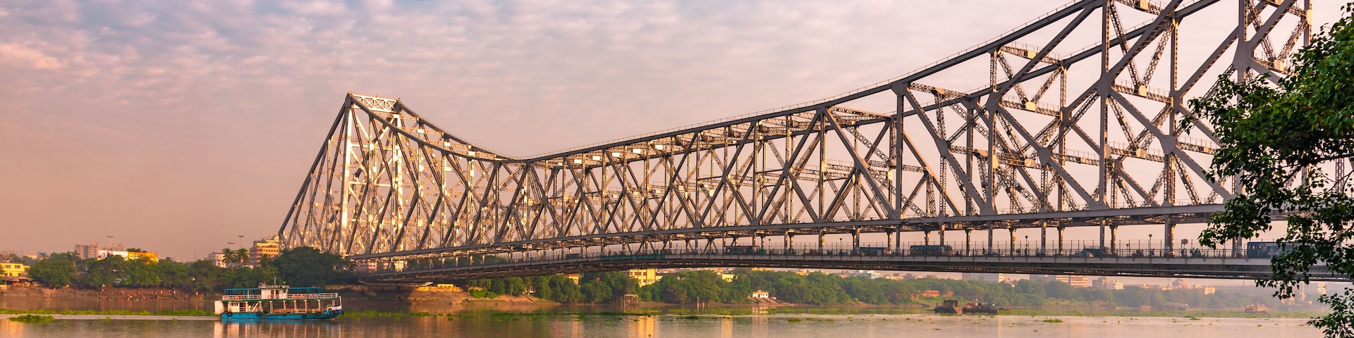 Un puente de acero grande que cruza un río, con una embarcación en primer plano y un paisaje urbano al fondo.