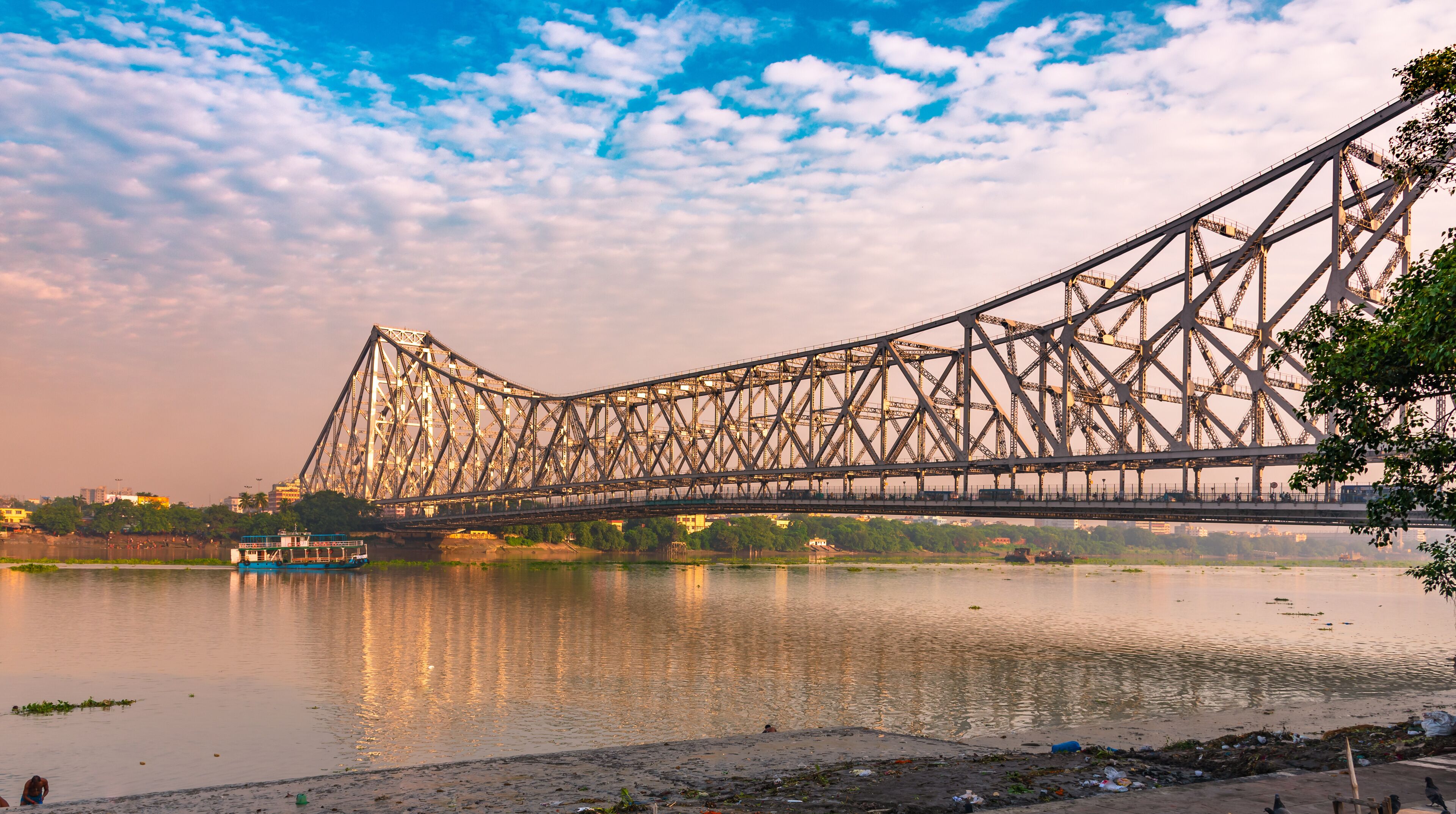 A large steel bridge spanning a river with a boat in the foreground and a cityscape in the background.