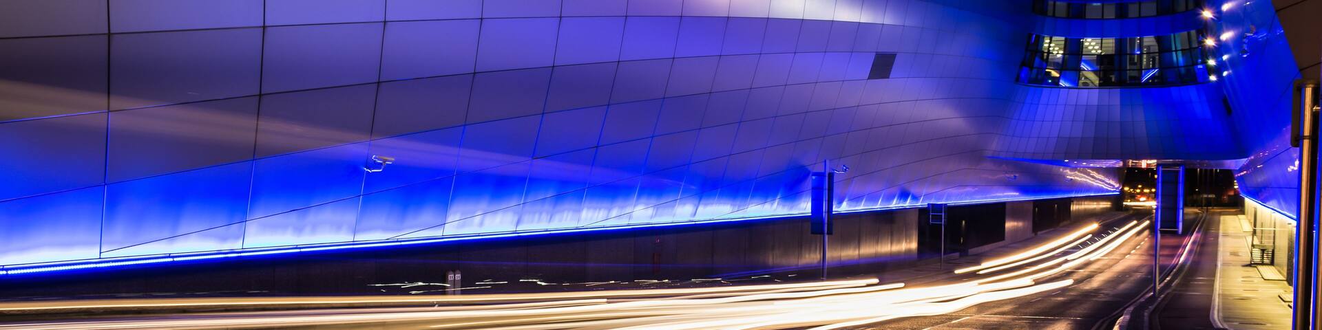 A nighttime urban street with light trails from moving vehicles, a modern building with blue lighting, and a curved structure with illuminated windows.
